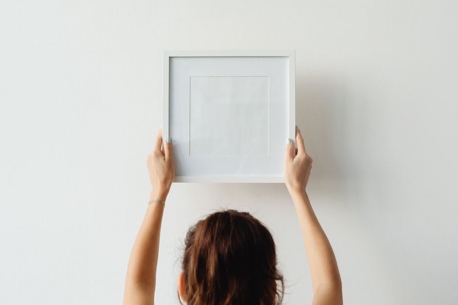 A person hanging a framed picture on an apartment wall using a level and screwdriver.