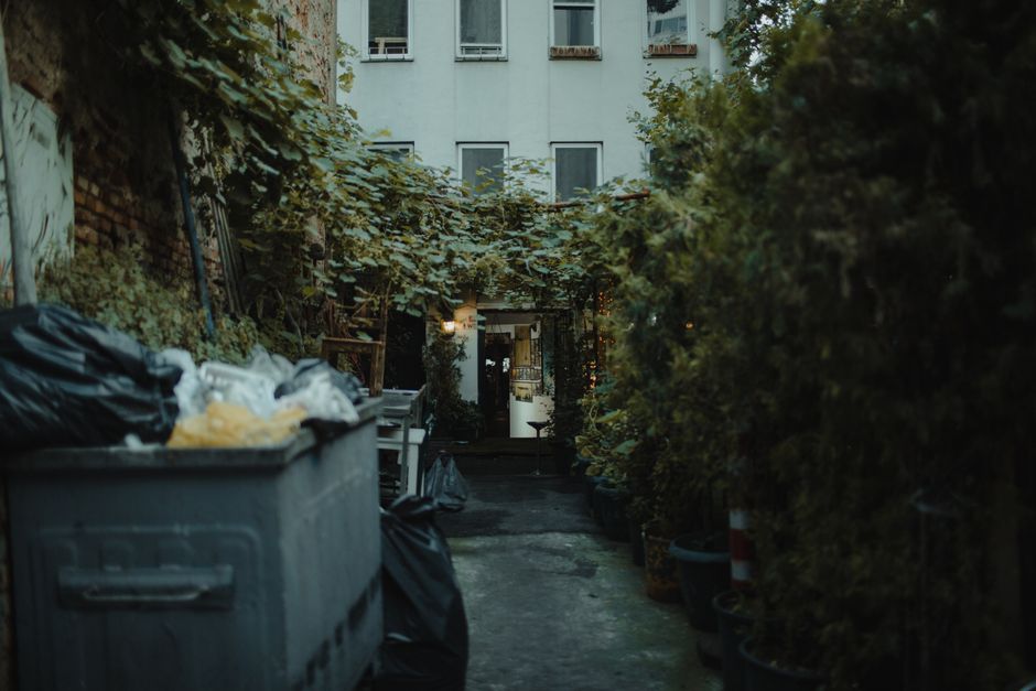 A shared apartment building trash room with tied garbage bags and bins lined against the wall